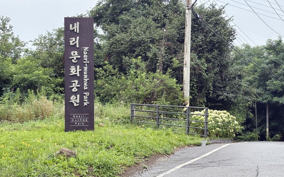 A dark brown wooden sign with Korean text stands beside a paved road. The sign is surrounded by abundant green vegetation and trees. The sign reads Naeri-munhwa Park in Hangul with English translation below.