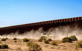 A military vehicle rolls along a dusty road at the border wall.