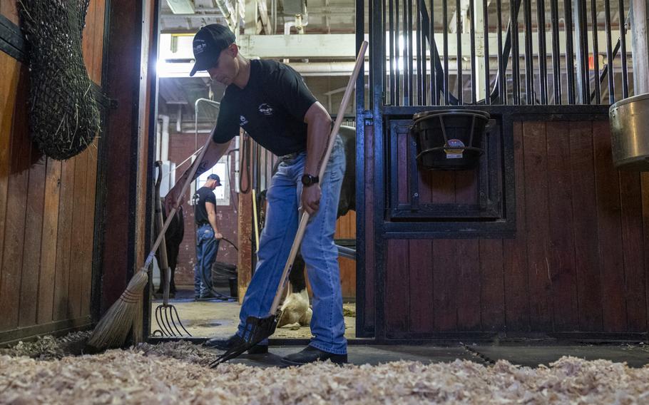 A soldier uses a broom to clean a stall. A rake rests against the wall.