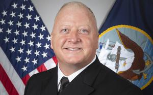 A naval officer in dress uniform poses for a portrait in front of U.S. and Navy flags.