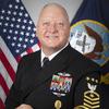 A naval officer in dress uniform poses for a portrait in front of U.S. and Navy flags.