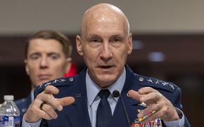 A seated Air Force general in blue dress uniform gestures with his hands while speaking into a microphone in a congressional hearing room.