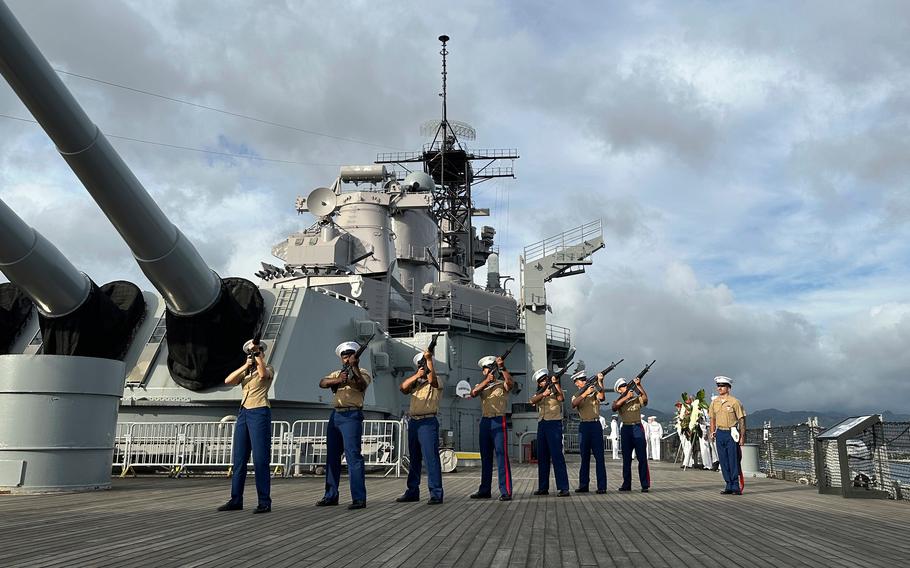 Marines in dress uniforms perform a rifle salute on the deck of a battleship, with the ship’s bridge behind them.