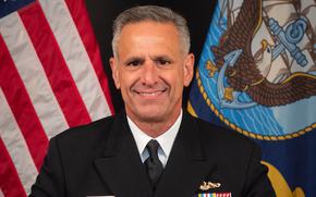 A U.S. Navy officer in dress uniform, seen from the chest up, poses in front of U.S. and Navy flags.