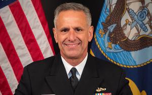A U.S. Navy officer in dress uniform, seen from the chest up, poses in front of U.S. and Navy flags.