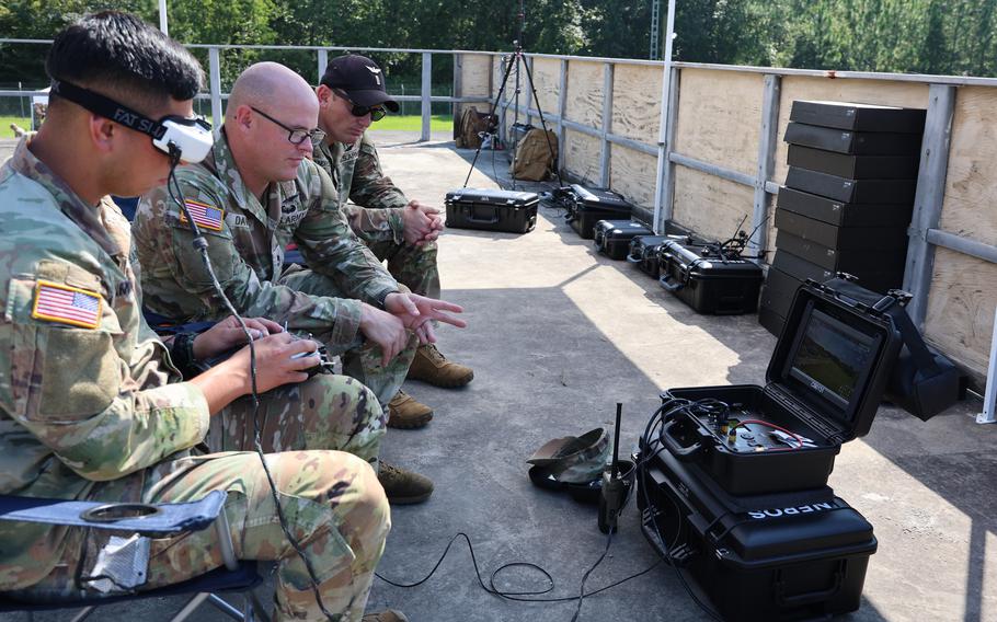 Soldiers seated outdoors, with one operating a remote control and wearing a VR headset.