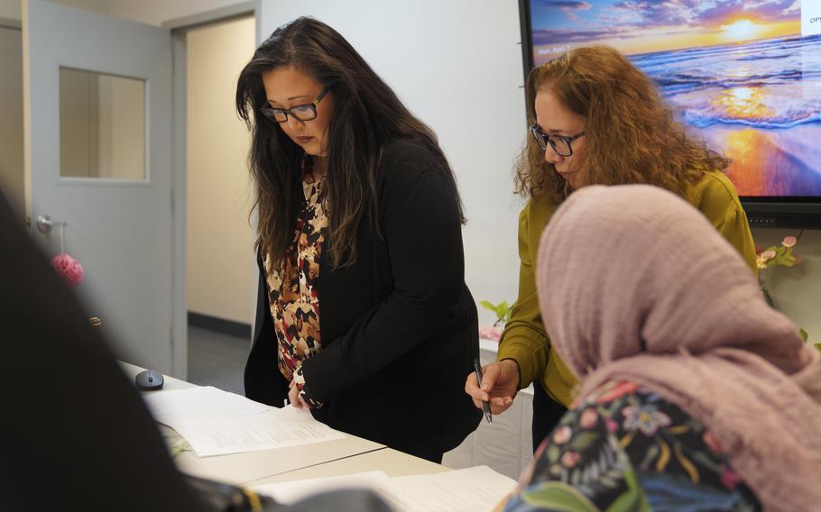 Two women in dark-rimmed glasses stand at a table in a conference room reading documents while a woman wearing a head scarf sits next to them in the foreground.