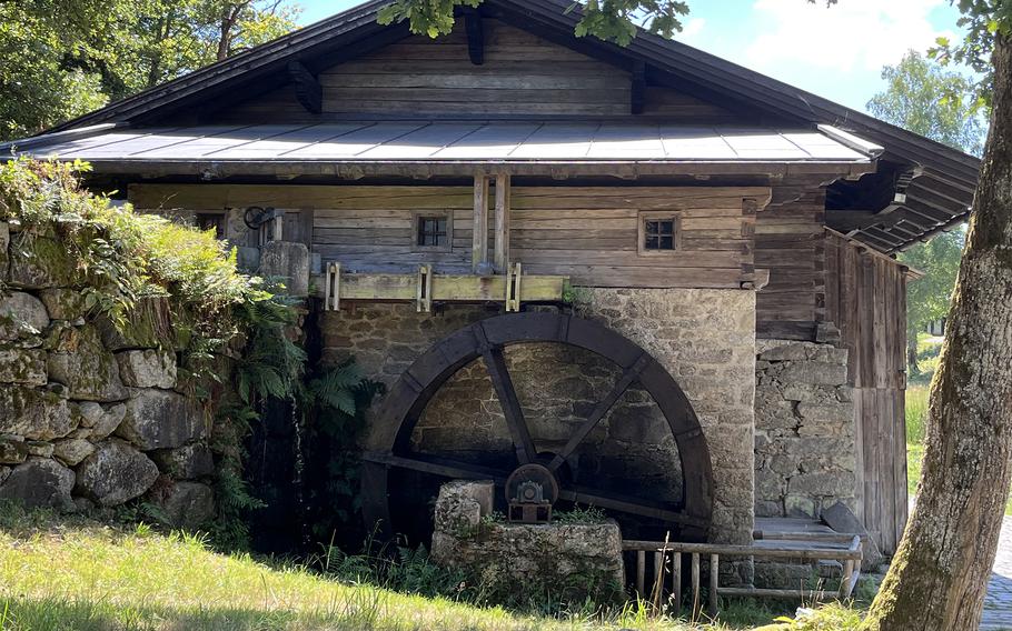 Exterior view of a traditional water mill with large wooden wheel visible through stone archway, surrounded by stone walls and wooden building structures with metal roofing.
