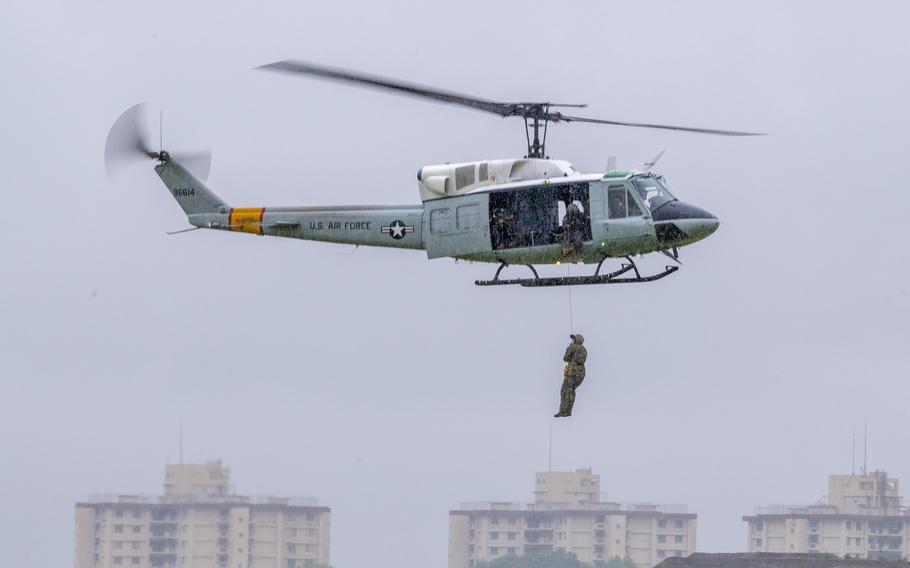 An airman in camouflage combat gear dangles on a line below a helicopter as it flies through the air with a city skyline in the background.