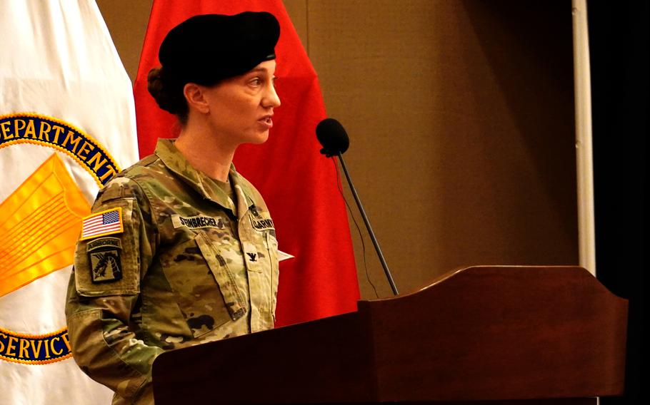 A woman in camouflage uniform and black beret stands at a wooden podium and speaks into a microphone.