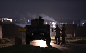 Silhouettes of armed soldiers and a military vehicle are seen at night on a dimly lit road. One soldier is positioned on top of the vehicle, while two others stand nearby.