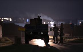 Silhouettes of armed soldiers and a military vehicle are seen at night on a dimly lit road. One soldier is positioned on top of the vehicle, while two others stand nearby.