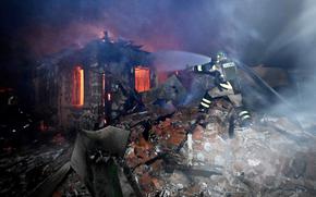 A firefighter stands on a pile of rubble and uses a hose to spray water on a burning building as smoke swirls above the scene.