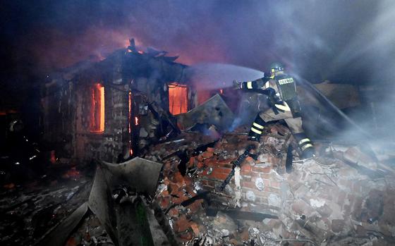 A firefighter stands on a pile of rubble and uses a hose to spray water on a burning building as smoke swirls above the scene.