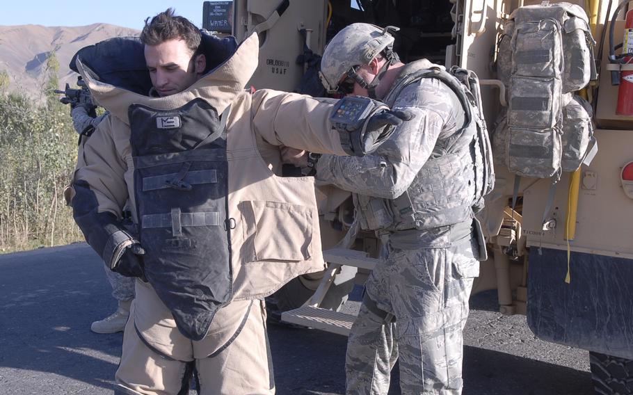 One service member helps another get outfitted in a bomb suit next to a military vehicle.
