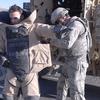 One service member helps another get outfitted in a bomb suit next to a military vehicle.