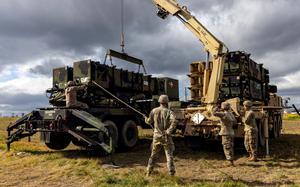 Military personnel in camouflage uniforms operate a yellow crane truck to lift and position green military equipment modules in an open field. 
