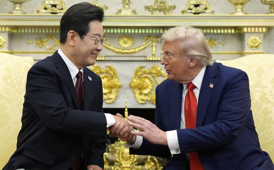 Donald Trump and South Korean President Lee Jae Myung, both wearing dark suits, lean towards each other and shake hands while seated in yellow chairs in front of a fireplace in the Oval Office of the White House.