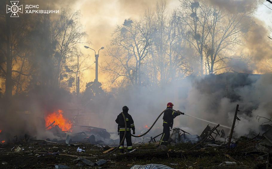 Two firefighters hold and spray a hose amid wreckage and fires.