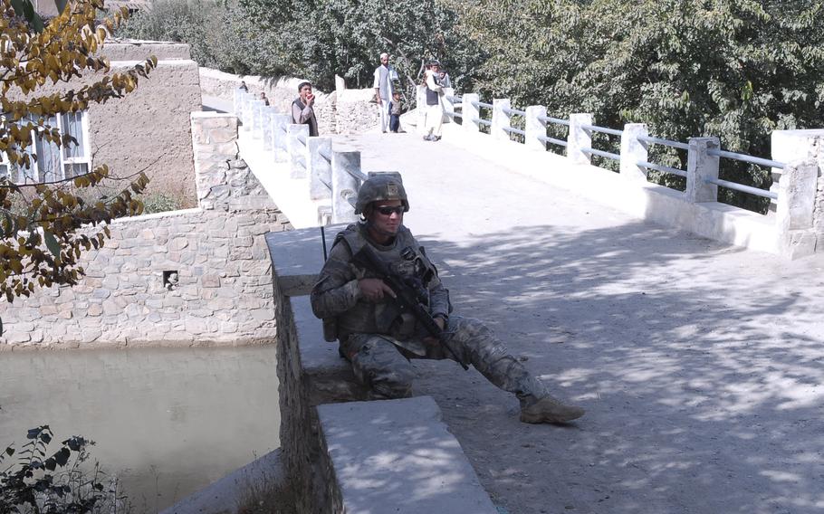 A soldier holding a rifle sits on a bridge as civilians stand behind him farther along the bridge.