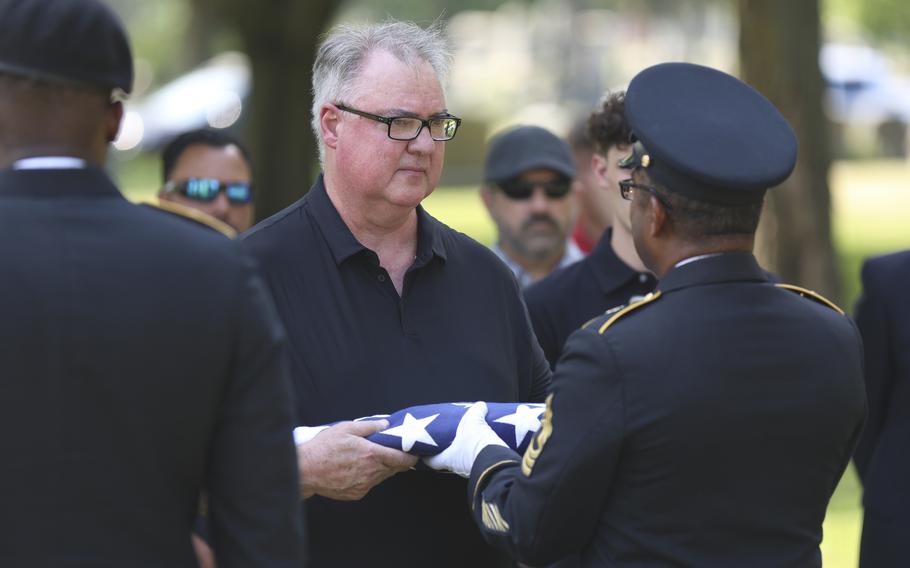A military officer in dress uniform and hat, seen from behind, presents a folded U.S. flag to a man in a dark polo shirt and dark-rimmed glasses during an outdoor funeral service.