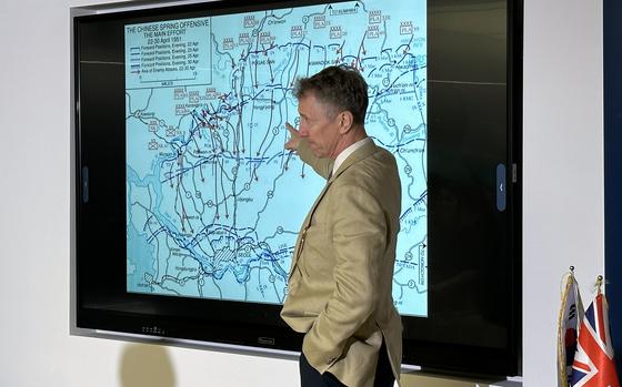 A man in a tan blazer stands before a large wall-mounted display showing a military map titled “The Chinese Spring Offensive — The Main Effort” dated April 1951, with various battle positions and movements marked. British and other flags are visible in the corner of the room.
