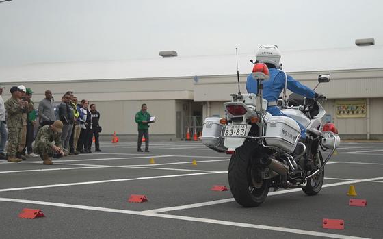 A police officer on a motorcycle, seen from behind, idles on an asphalt training course as a group of observors, including military members in camouflage uniforms, watches on the left.