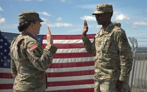 A soldier raises their right hand while taking an oath in front of another soldier, who is administering the oath. Both are dressed in camouflage uniforms, standing outdoors with a large American flag displayed behind them. 