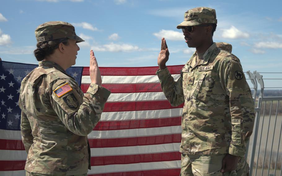 A soldier raises their right hand while taking an oath in front of another soldier, who is administering the oath. Both are dressed in camouflage uniforms, standing outdoors with a large American flag displayed behind them. 