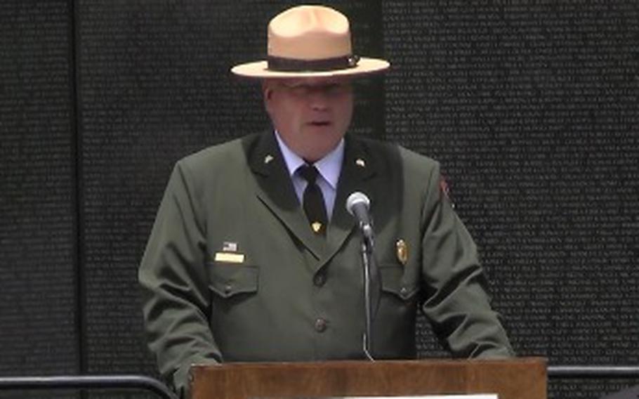 A man in a green park ranger uniform and tan hat stands at a podium and speaks into a microphone in front of a black marble wall engraved with the names of troops killed in war.