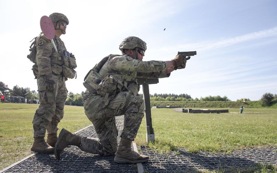 One soldier stands and watches as another soldier kneels and fires a hand gun.