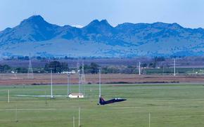 A blue fighter jet takes off in a rural landscape with mountains in the background.