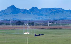 A blue fighter jet takes off in a rural landscape with mountains in the background.