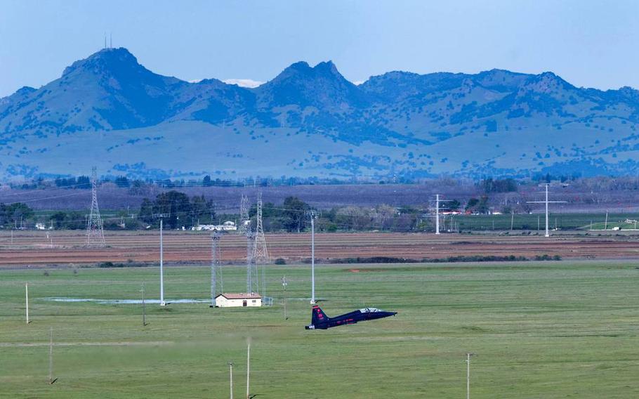 A blue fighter jet takes off in a rural landscape with mountains in the background.