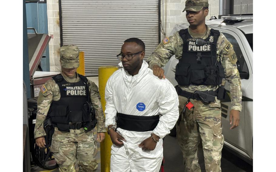 A man in a white jump suit is escorted by two soldiers with military police vests.