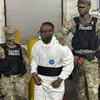 A man in a white jump suit is escorted by two soldiers with military police vests.