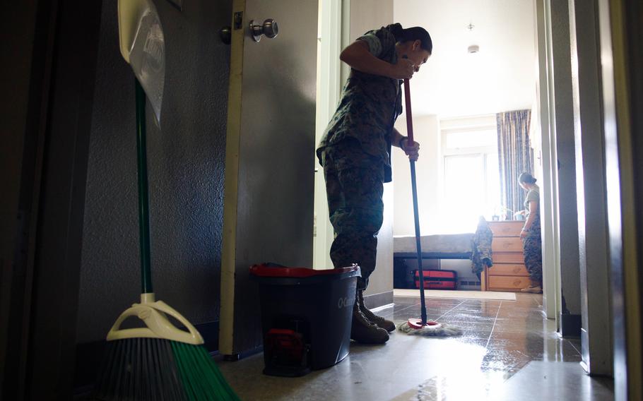 Two women in fatigues clean a room. One is mopping, the other standing next to a dresser in the background.