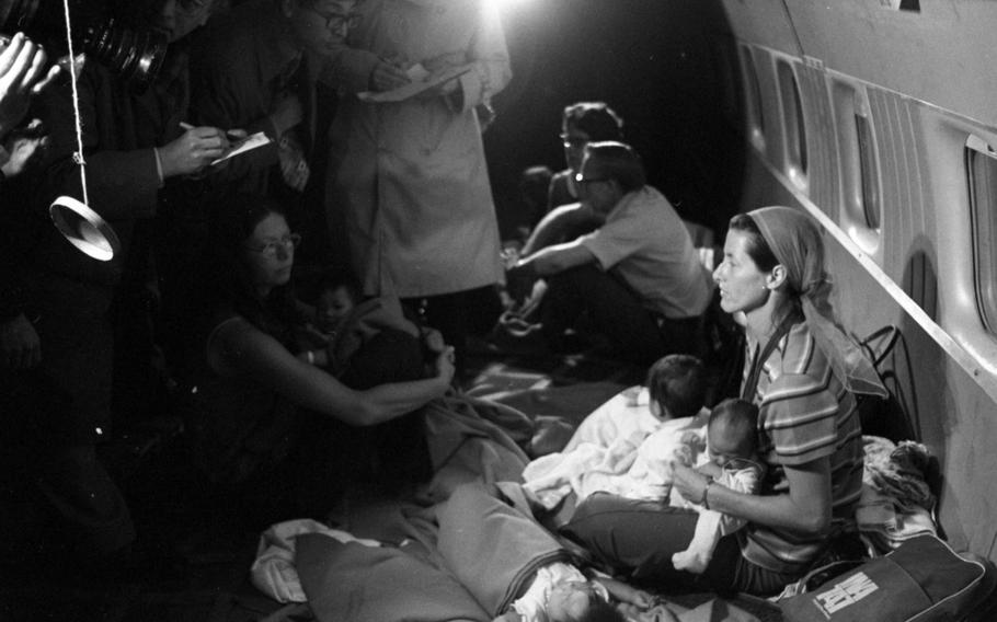 A black-and-white, 1970s-era photo of a woman seated on the floor of an airplance holding two babies as reporters stand in front of her.