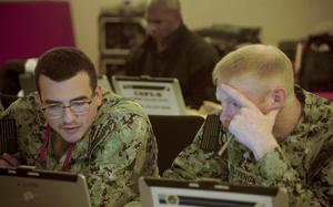 Two service members leaning over computers while seated.