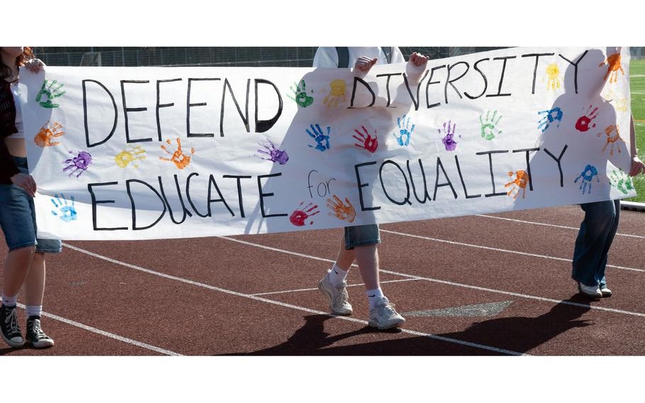 Three students hold a large banner that reads “DEFEND DIVERSITY” and “EDUCATE for EQUALITY.”
