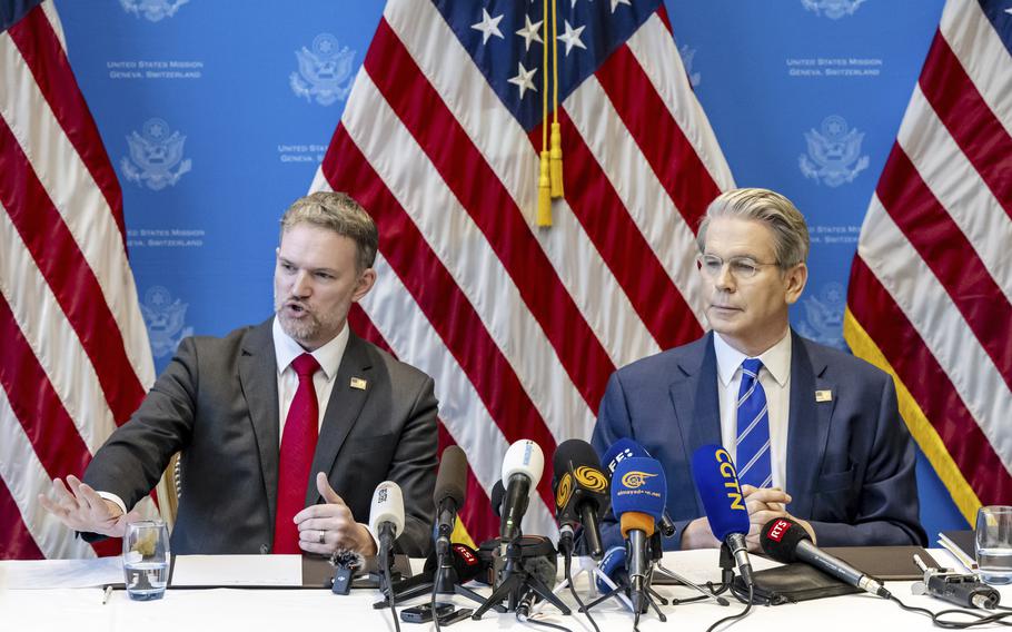 Two government officials in suits sit at a table with microphones in front of large U.S. flags.