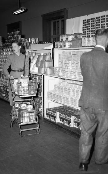 A vertical, black and white photo of two people at a commissary; one is a man seen from the back, the other is a woman with a shopping cart.