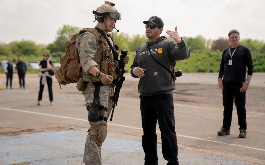 Two men in the center, one in military fatigues and the other in a hoodie and pants, with four others standing out-of-focus behind them.