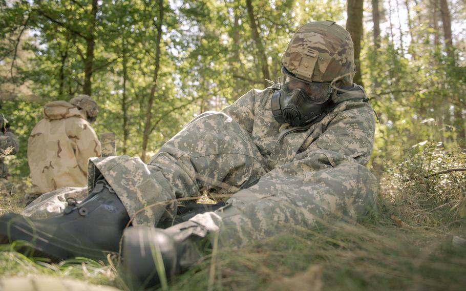A soldier in full gear and a gas mask sits on the ground with boots out.