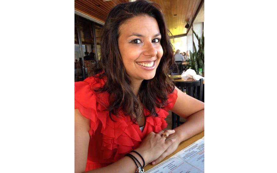 A close-up view of a woman in a red shirt smiling and posing as she sits at a table in a restaurant, with other tables in the background.