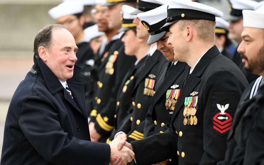 A man in an overcoat shakes hands with naval officers in dress uniforms and hats standing in a line.