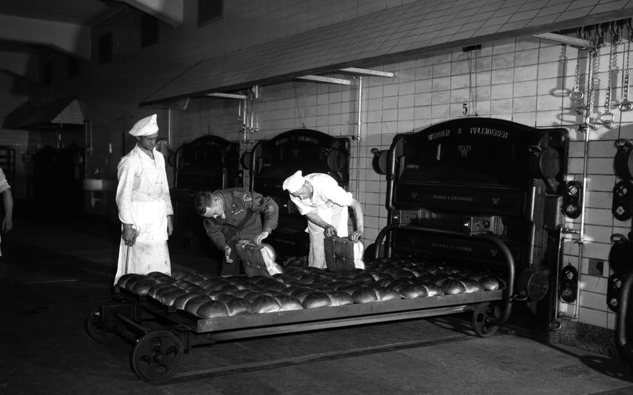A man in a service uniform inspects bread next to two men in white kitchen uniforms.