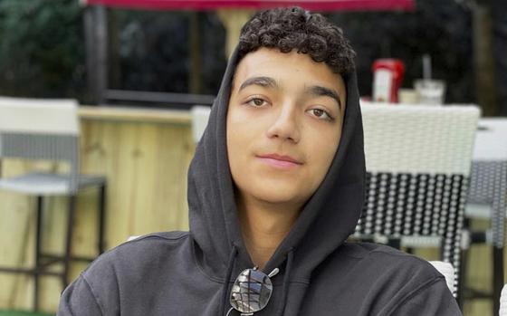 A boy wearing a black hoodie and sunglasses smiles slightly.