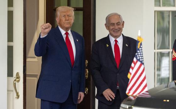 Trump raises his right hand and makes a fist, and Netanyahu stands next to him, smiling. An American flag is on the front of a car in front of them.