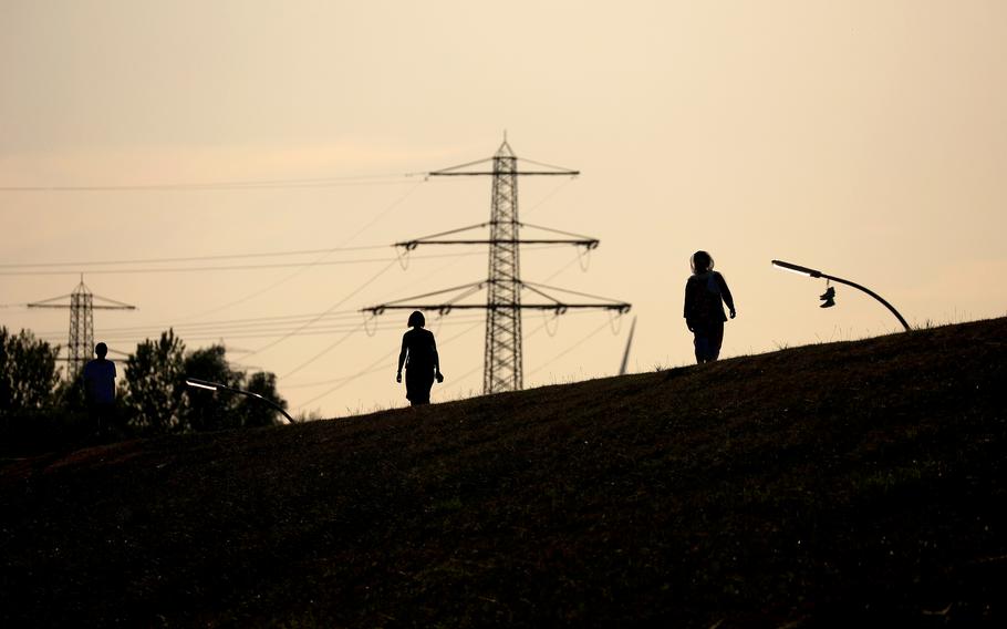 High voltage electricity towers beyond pedestrians on a river bank in Hamburg, Germany, on Aug. 24, 2022. 